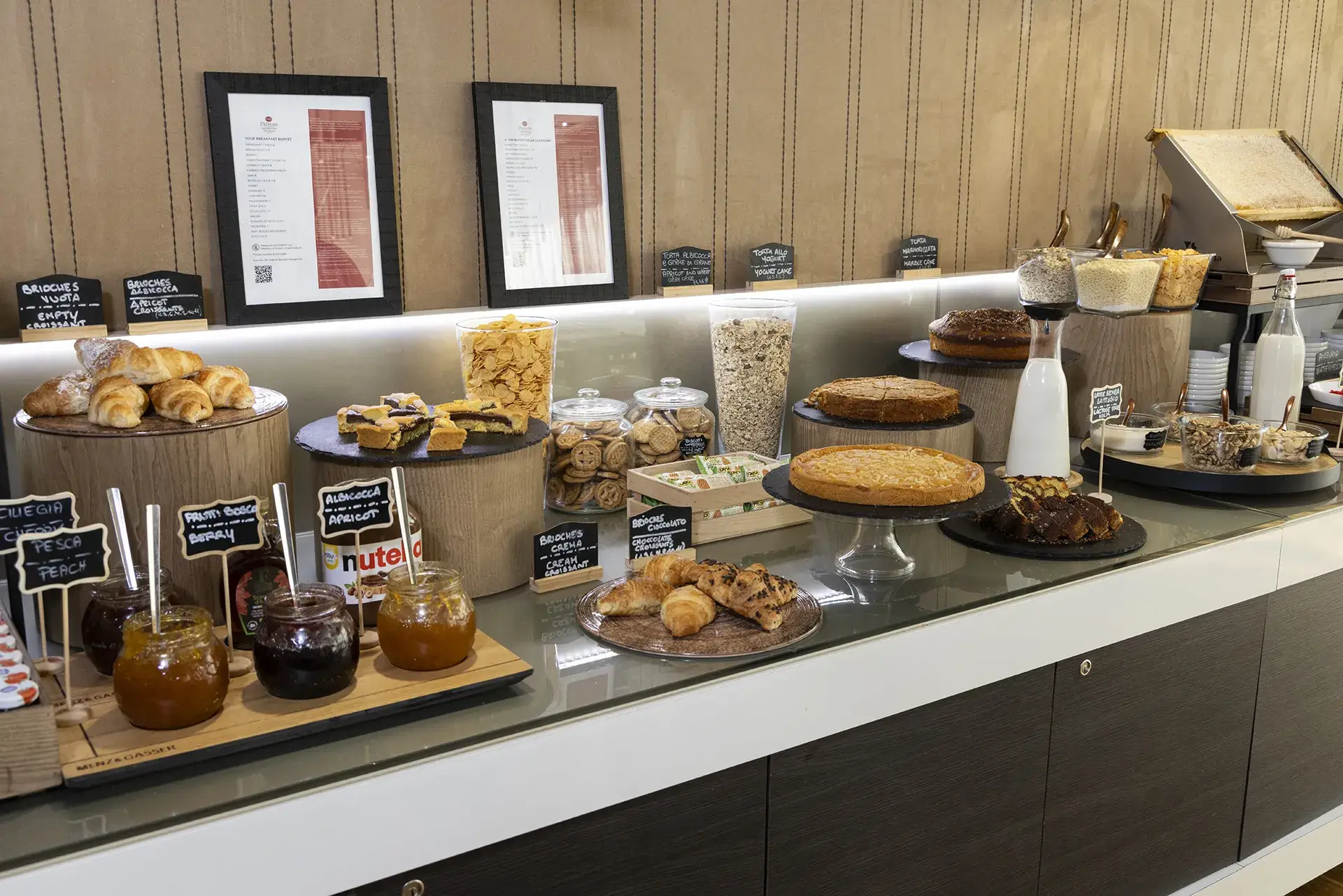 The image shows a portion of the hotel's breakfast buffet. On the left are open jars of jam, maple syrup, and Nutella. Behind and to the side are plates of croissants and sliced ​​cake. In the center are tall jars of granola and cereal, filled cookie jars, a tray of packaged cookies, and cake stands with various cakes. On the right is a jug of milk and containers of seeds, cereals, and granola.