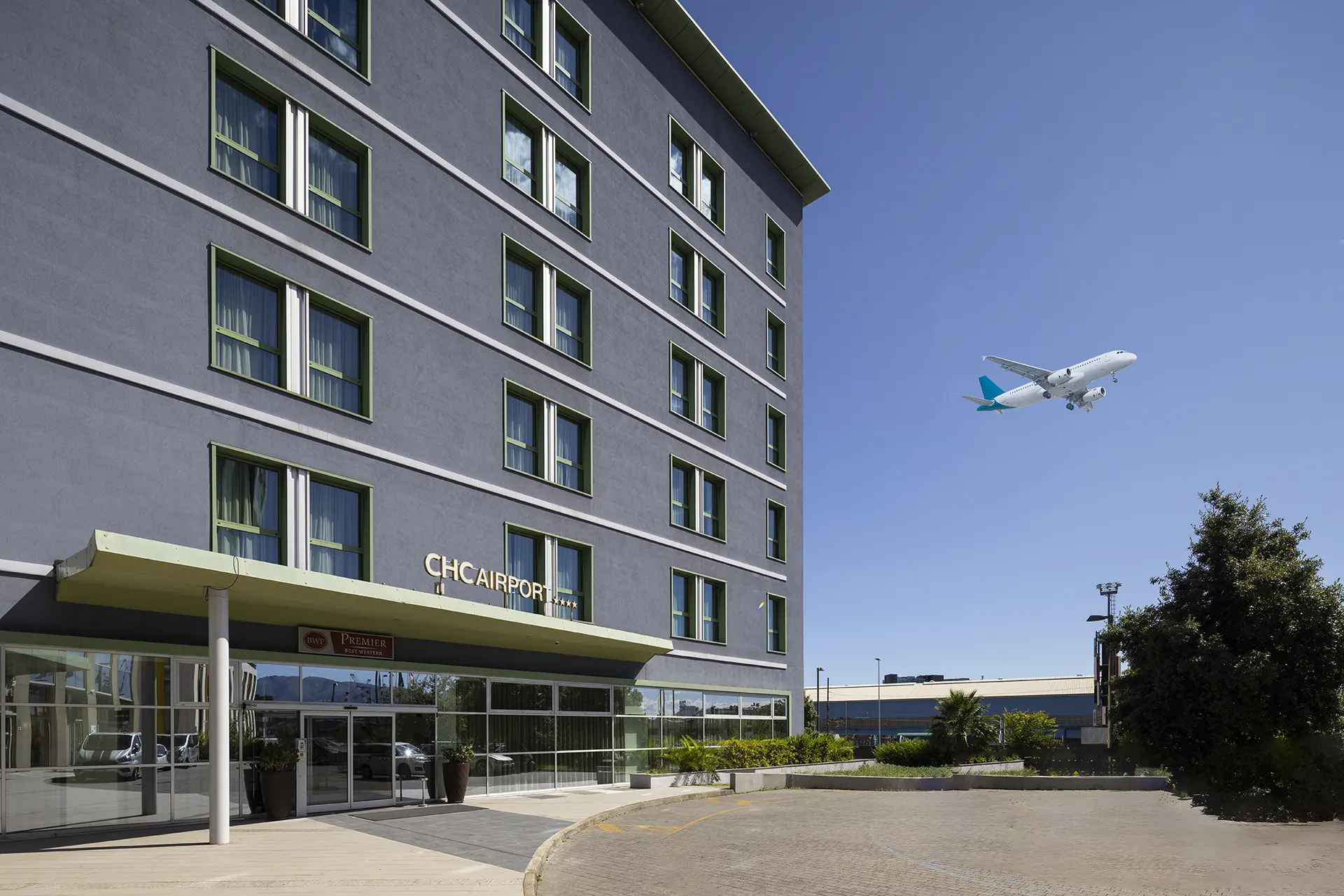 The image shows the outside of the hotel with a plane just taken off from the airport in the sky next to it.