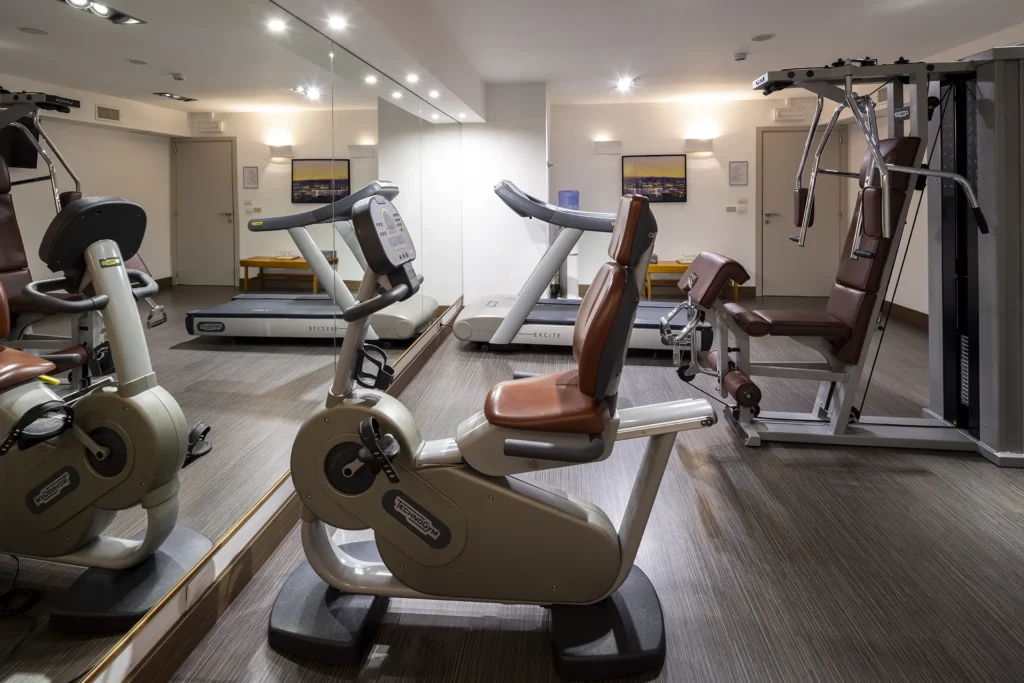 The photo shows the hotel's mini gym with various exercise equipment. There's an exercise bike, a multi-function bench, and a treadmill. The left wall is covered with mirrors.
