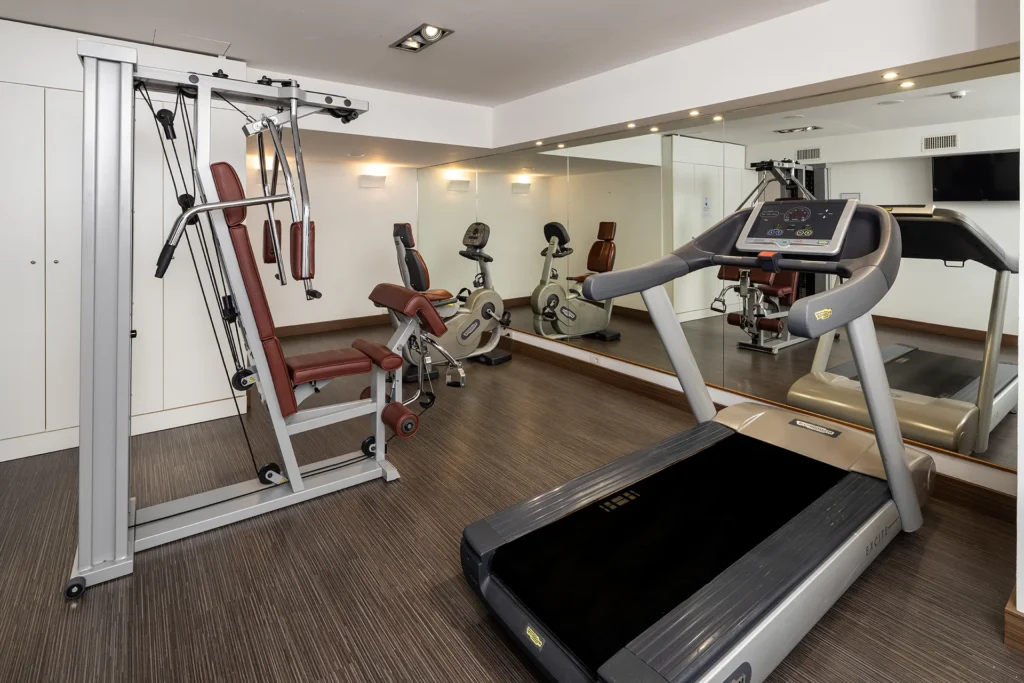The photo shows the hotel's mini gym with various exercise equipment. There's an exercise bike, a multi-function bench, and a treadmill. The right wall is covered with mirrors.